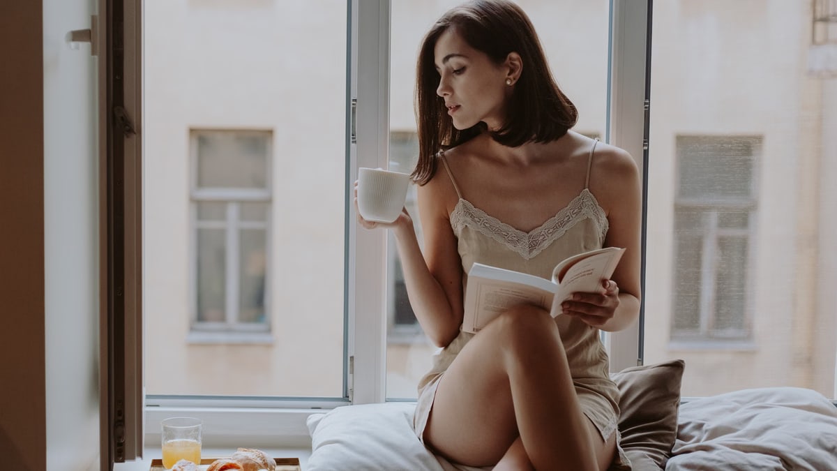 Woman in sleepwear enjoys coffee while reading by the window on a relaxed morning.